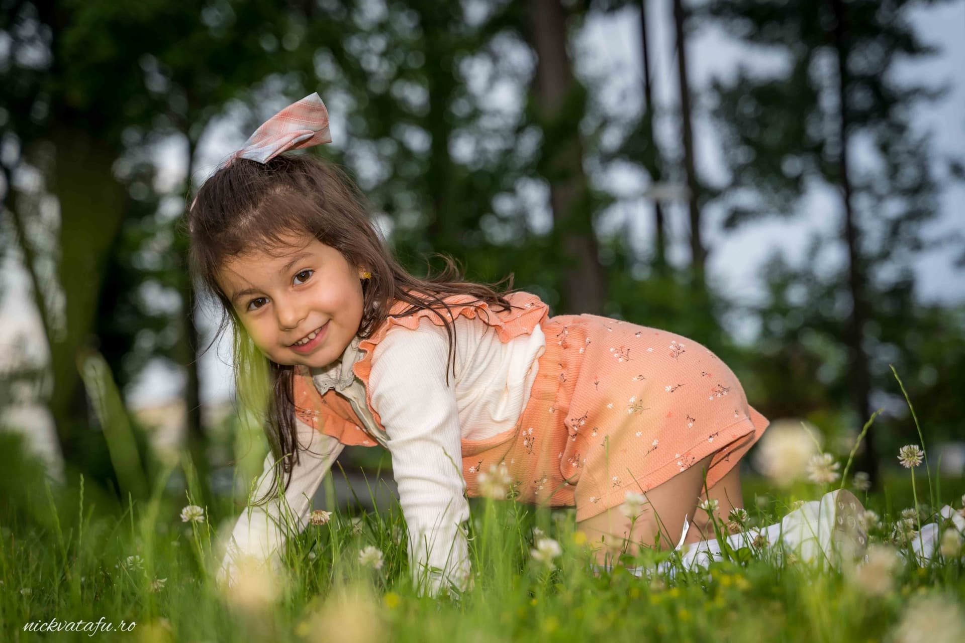 Fetiță zâmbitoare, cățărându-se pe iarbă, surprinsă într-un moment jucăuș și plin de emoție.