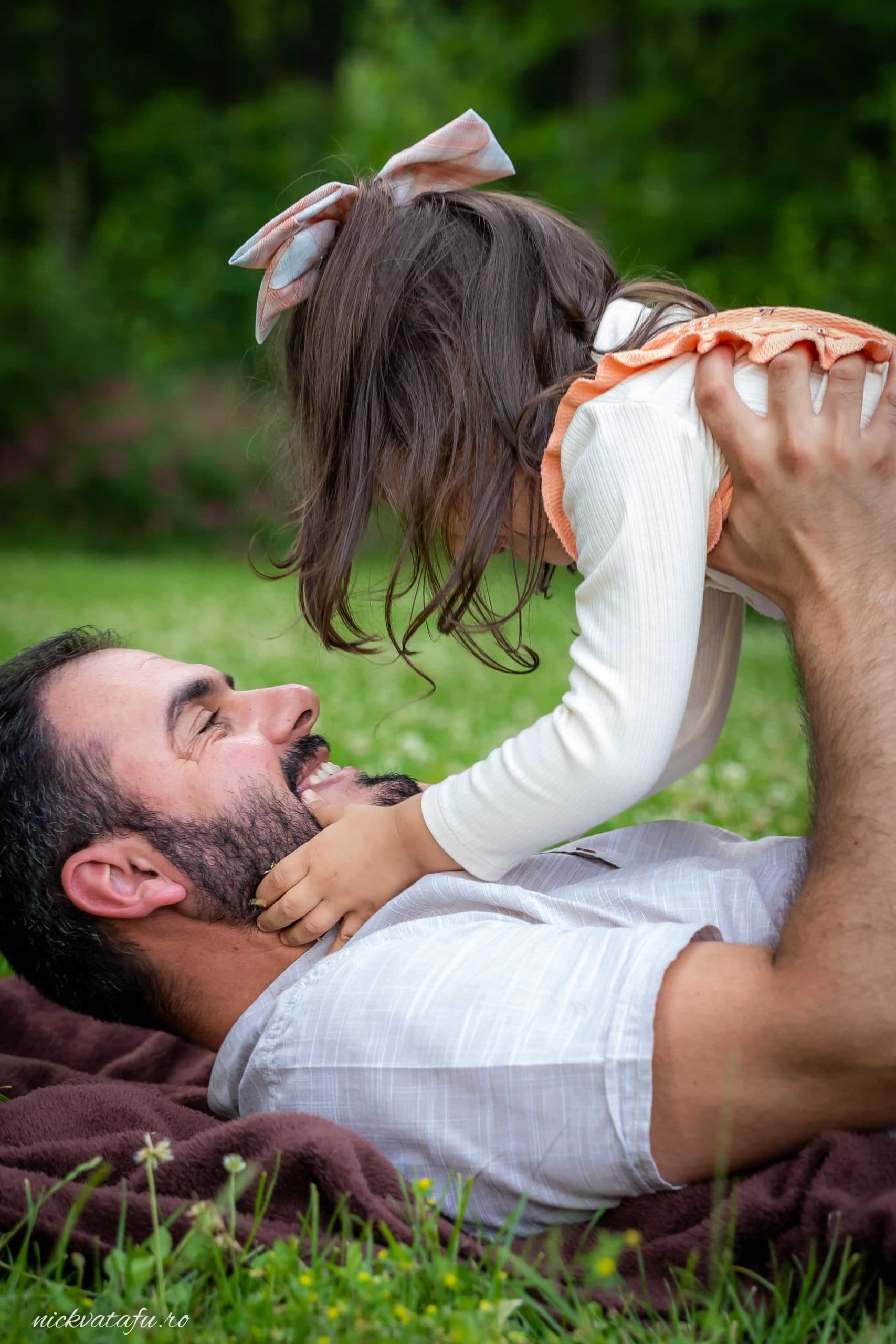 Bărbat zâmbitor se joacă cu o fetiță pe gazon, surprinzând emoția unei legături familiale.
