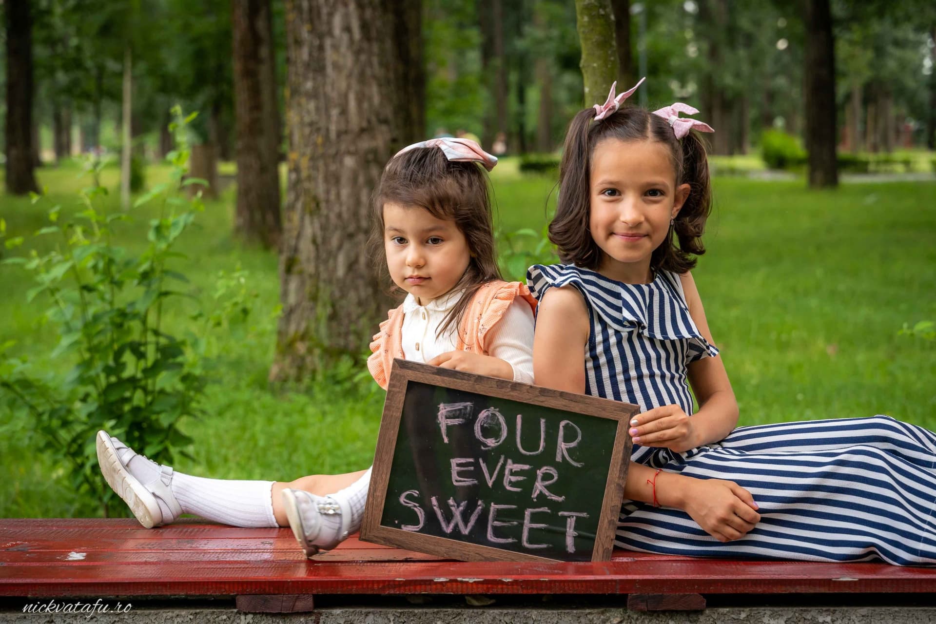 Două fetițe zâmbitoare stând pe o bancă, ținând un tablou cu mesajul „FOUR EVER SWEET,” într-un parc verde.