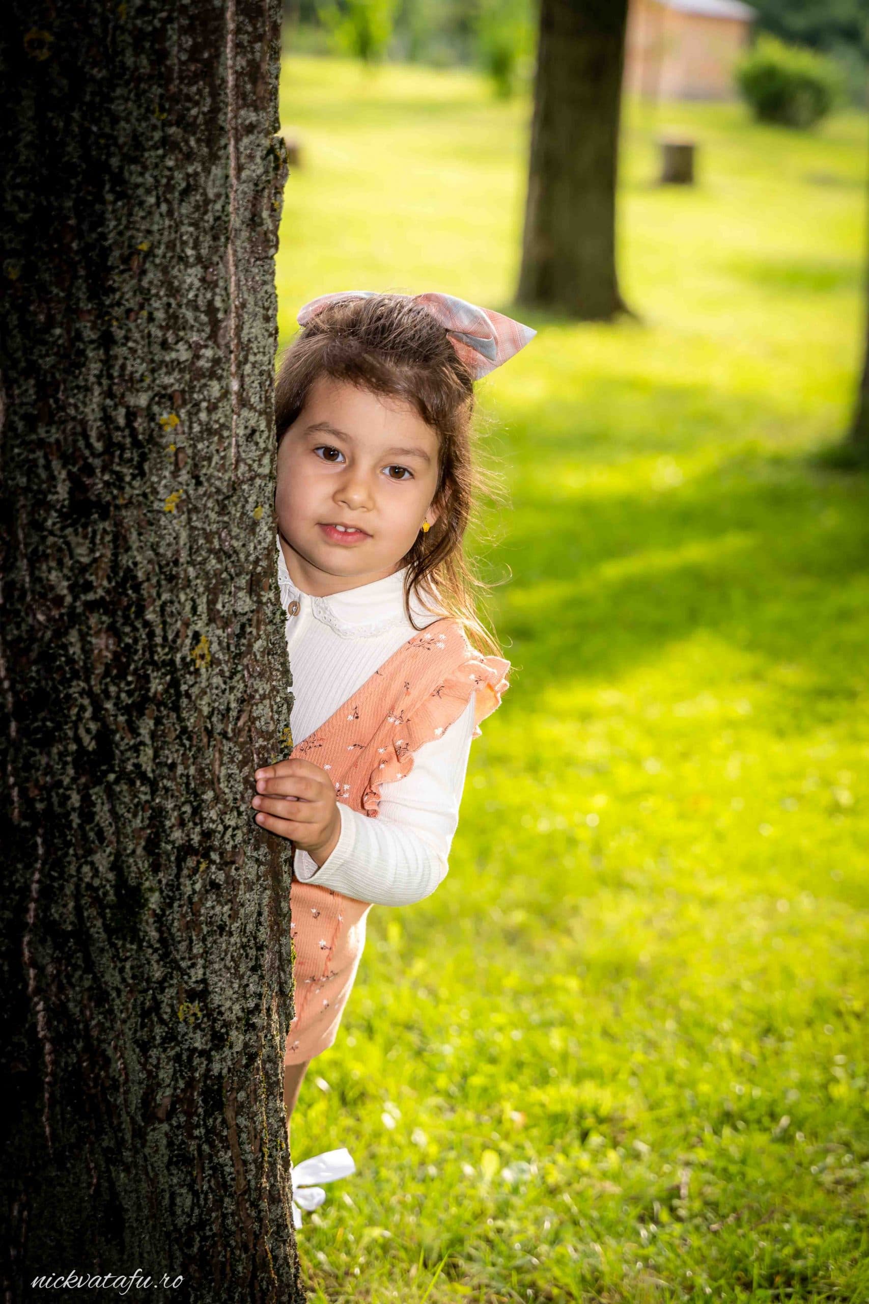 Fetiță jucăușă ascunzându-se în spatele unui copac, cu zâmbet și emoție, în cadrul unei fotografii de nuntă.