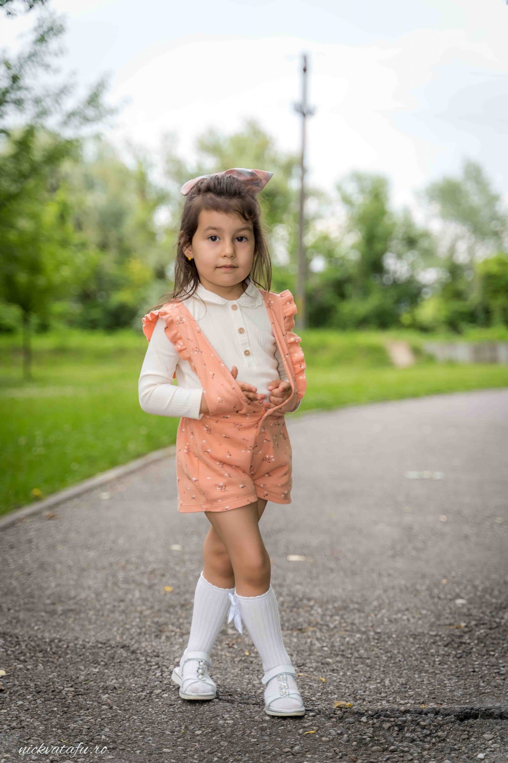 Fetiță zâmbitoare într-o ținută elegantă, pe aleea unui parc, evocând emoții de bucurie și inocență.