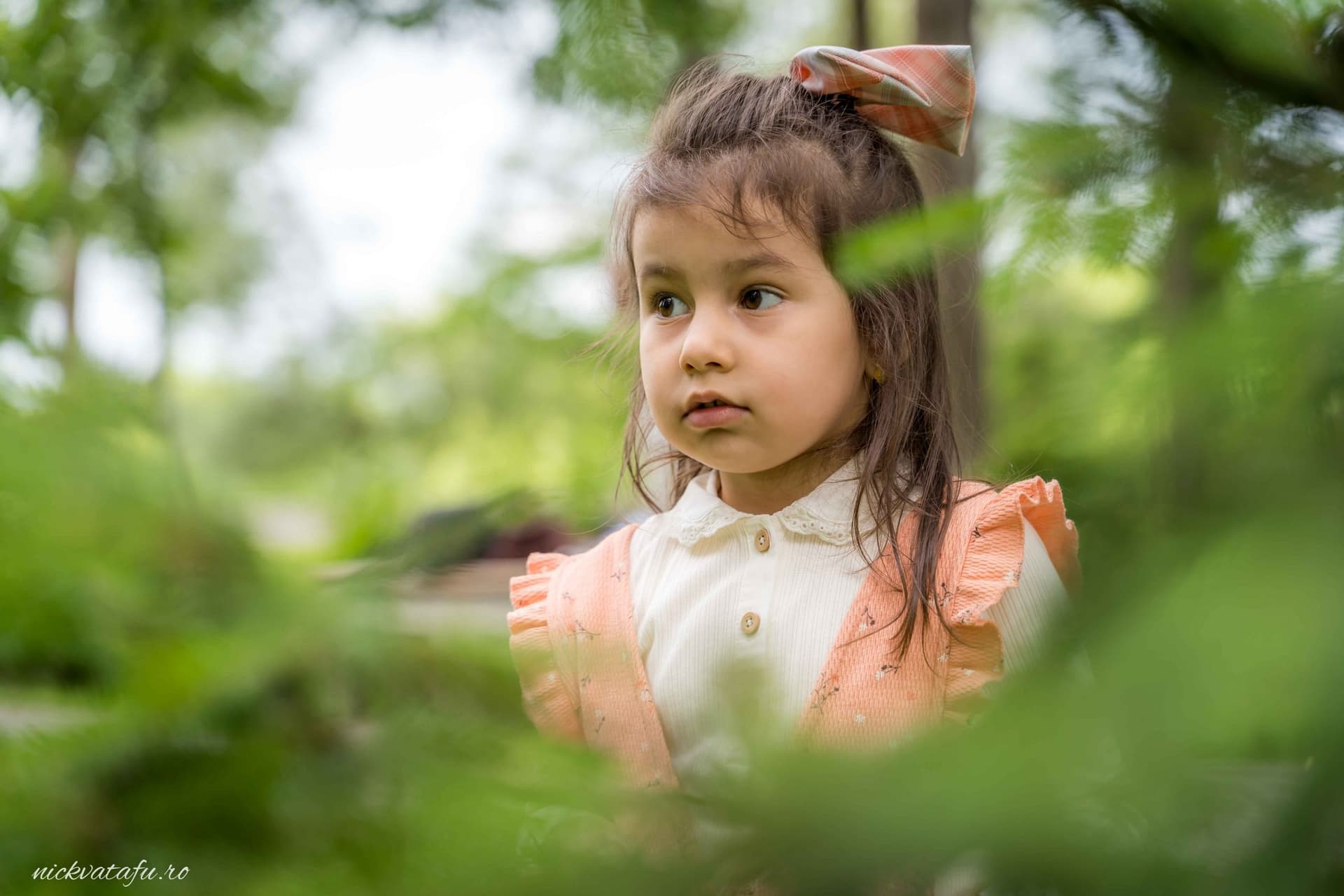 Fetiță cu o expresie curioasă, înconjurată de verdeață, surprinsă într-o fotografie de botez în natură.