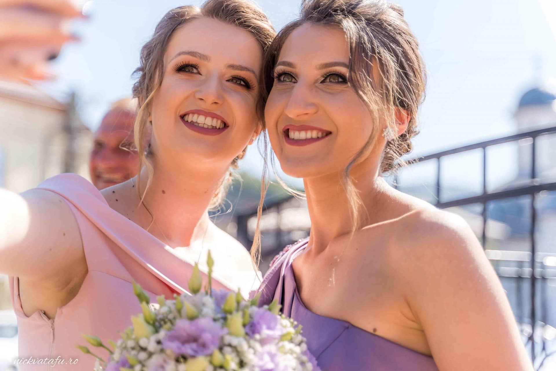 Două domnișoare de onoare zâmbesc fericite, purtând rochii elegante și buchete de flori, la o nuntă în aer liber.