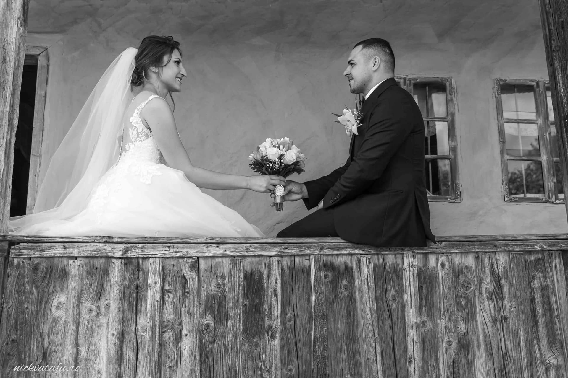 Mire și mireasă zâmbind unul către altul, ținând o floare, fotografie elegantă de nuntă în alb-negru.