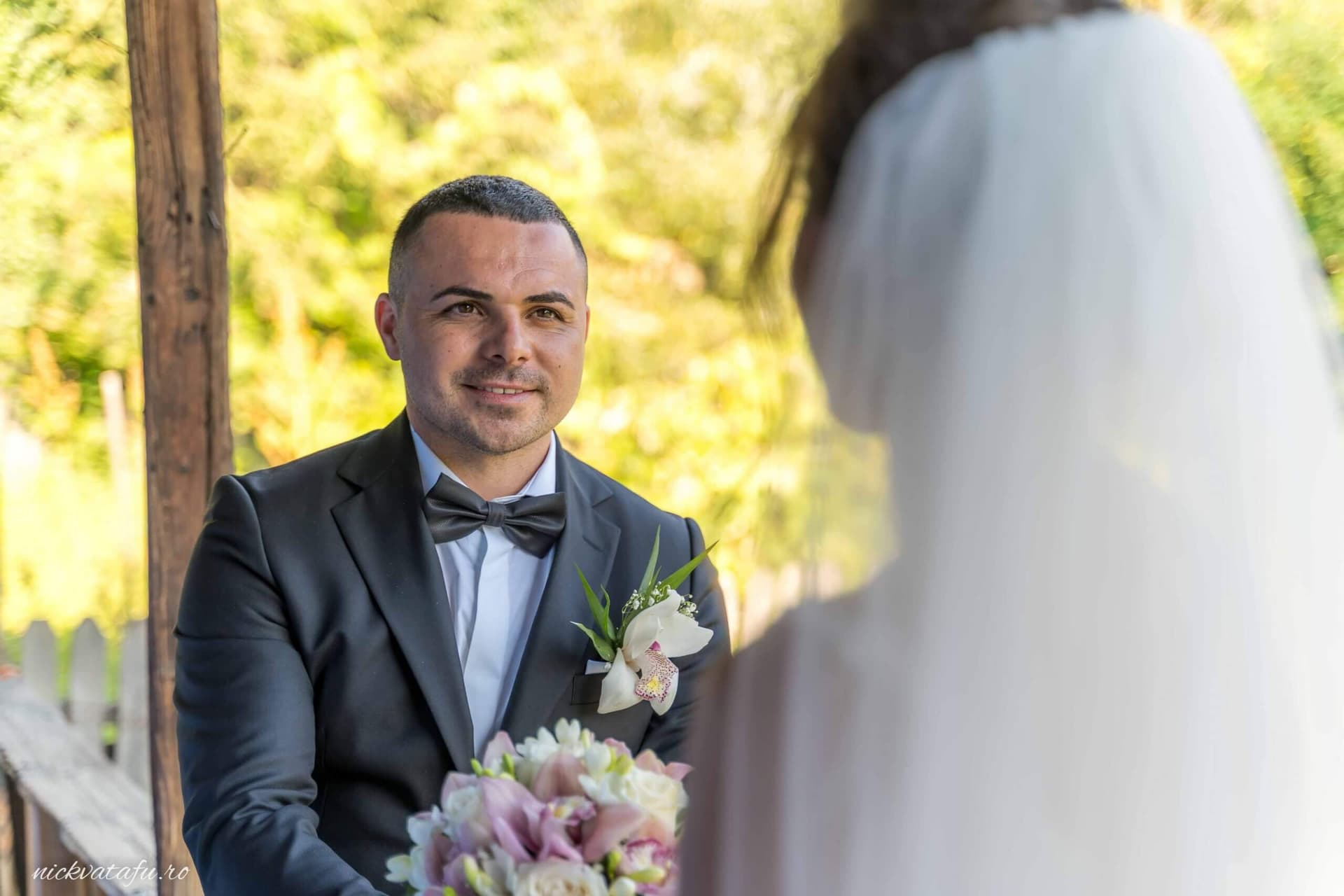 Mirele zâmbește emoționat către mireasă, în fundal verde, fotografie de nuntă plină de iubire și detalii delicate.