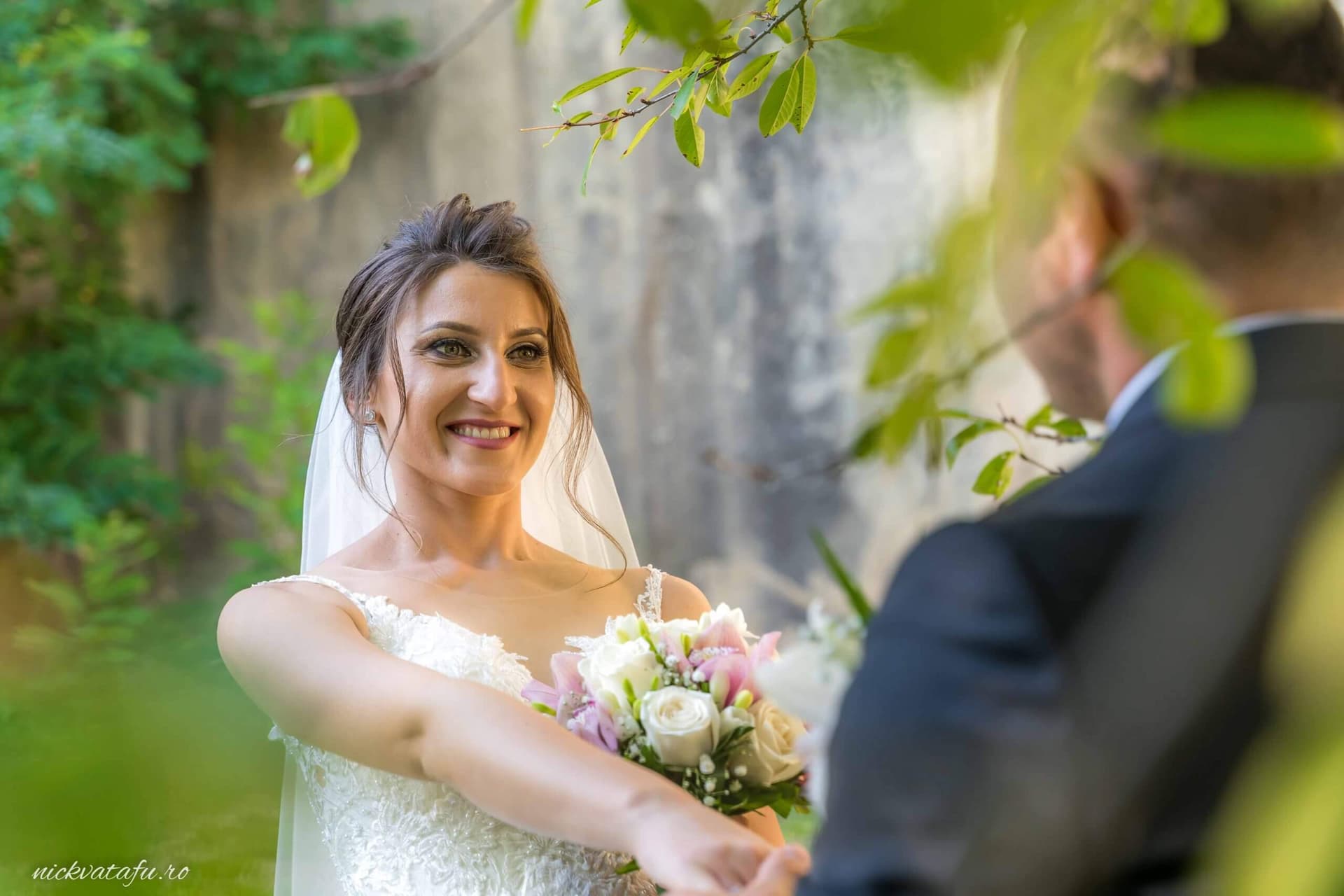 Mireasă zâmbitoare oferind buchetul soțului într-un cadru natural, fotografie nuntă plină de emoție.