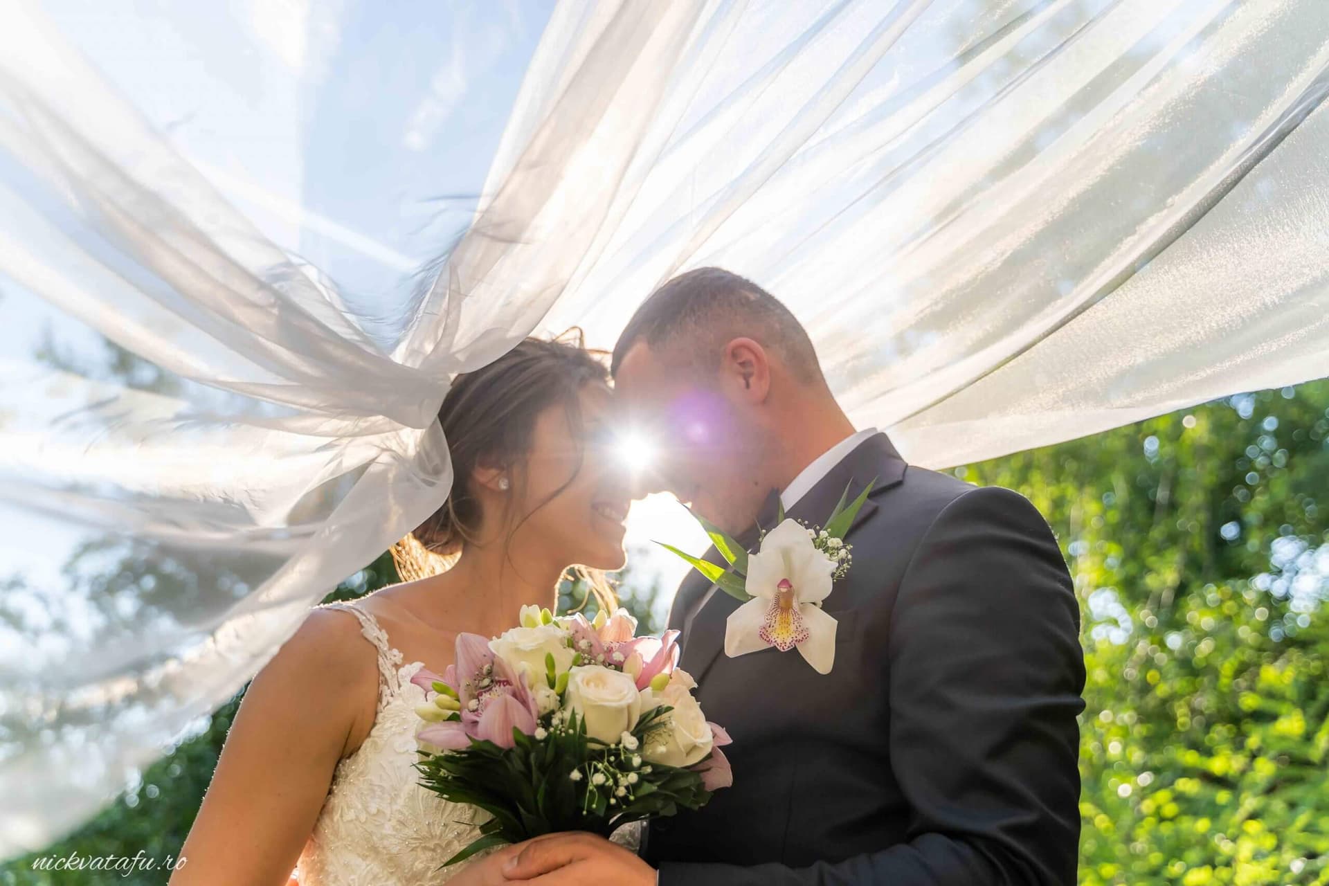 Mire și mireasă îmbrățișați sub voal, cu razele soarelui strălucind, fotografie de nuntă plină de emoție.
