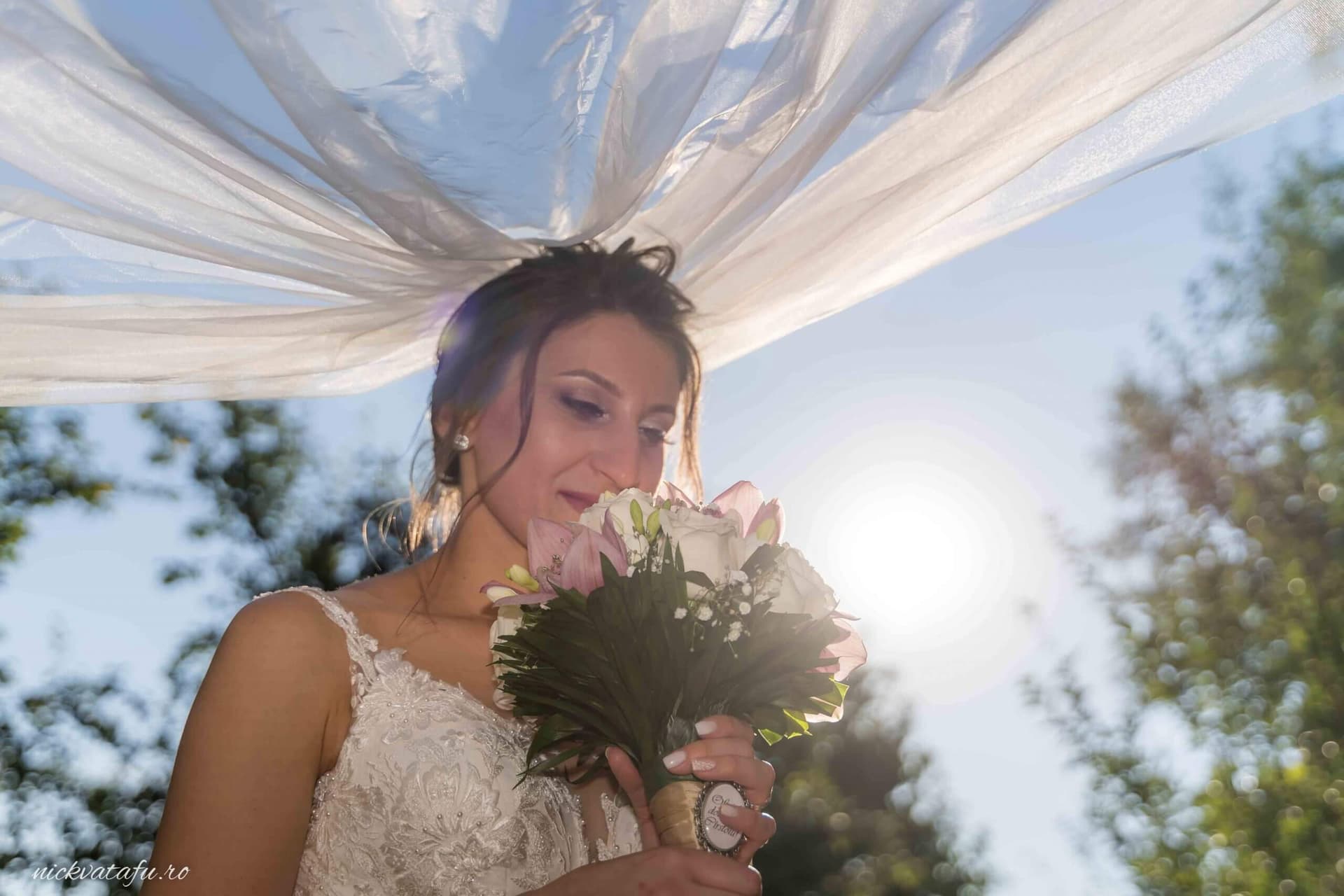 Mireasă emoționată, cu buchet de flori, sub voal, în lumina blândă a soarelui, fotografie nuntă elegantă.
