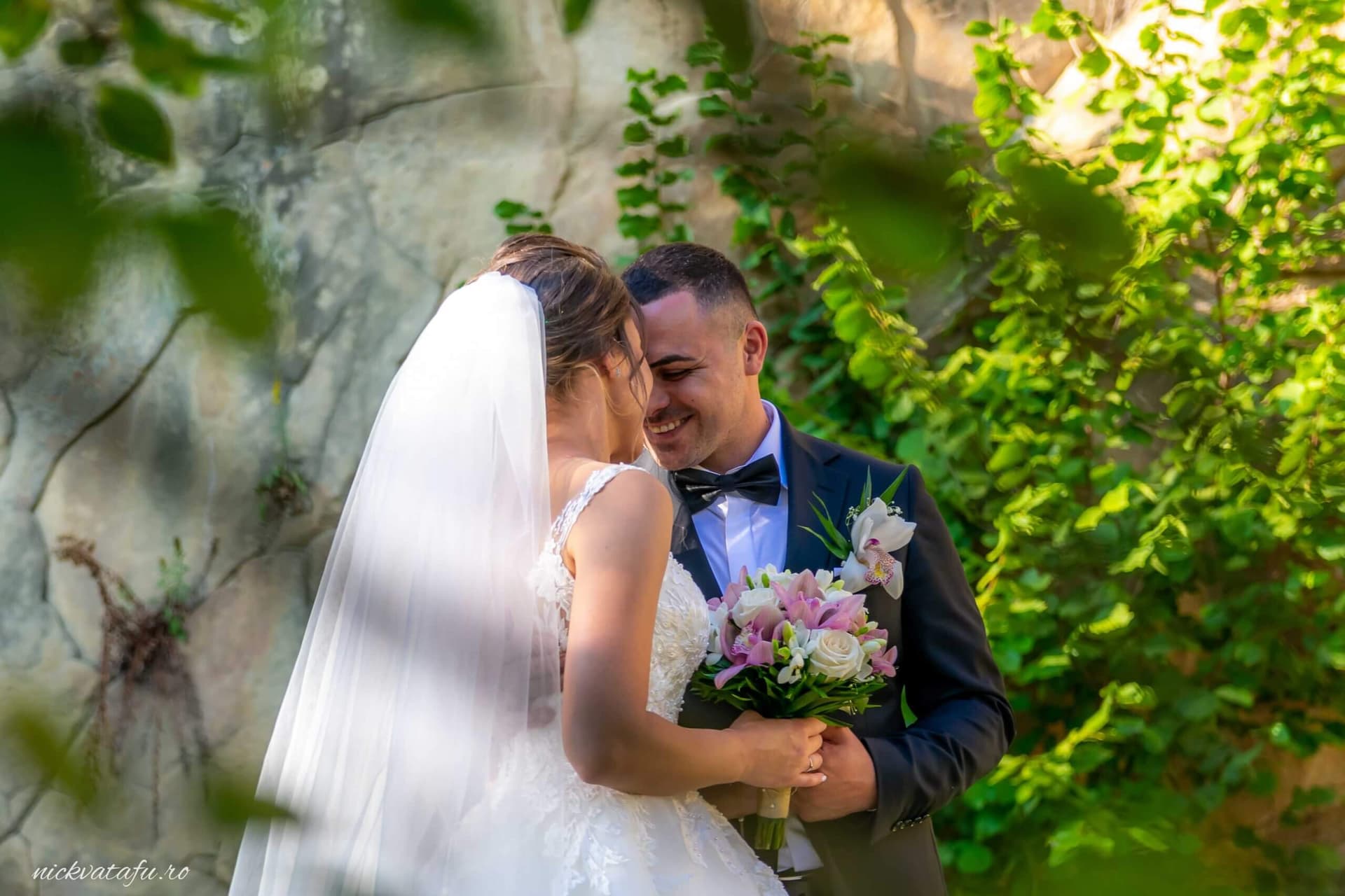 Mire și mireasă zâmbind unul către altul, înconjurați de verdeață, fotografie de nuntă plină de emoție.