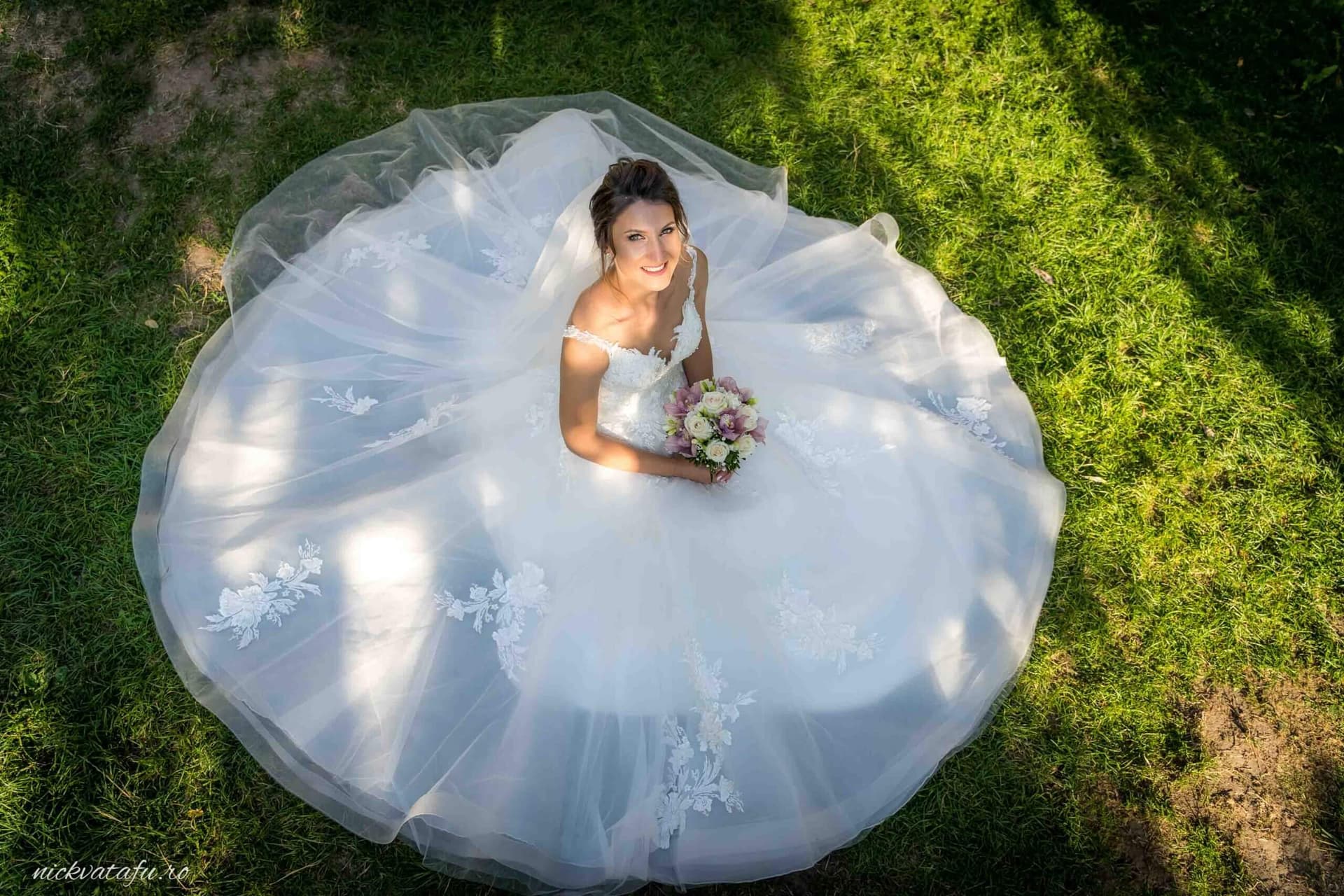 Mireasă zâmbitoare, în rochie albă, stând pe iarbă, cu buchet în mână, fotografie nuntă elegantă.