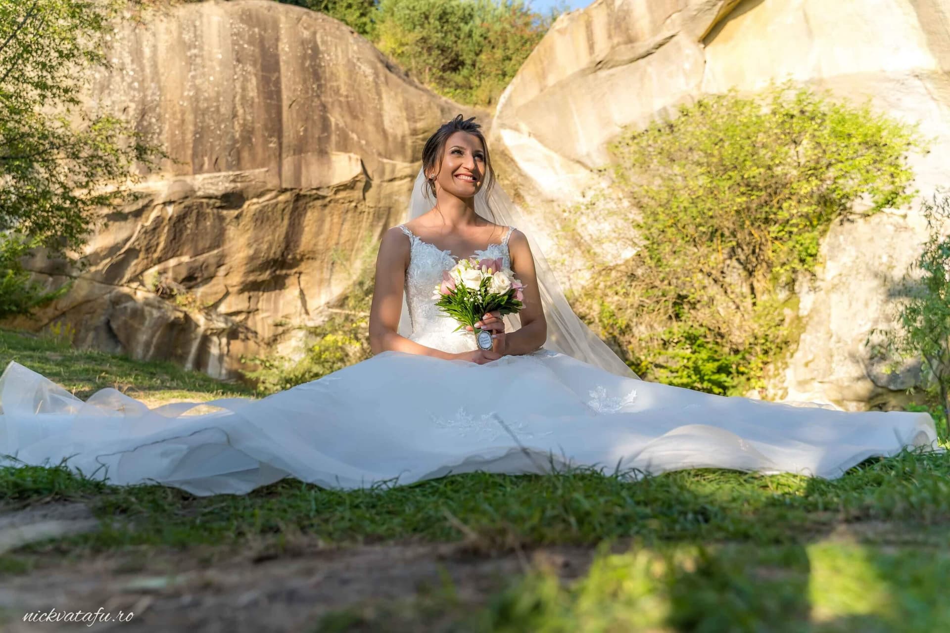 Mireasă zâmbitoare, așezată pe iarbă, înconjurată de natură, fotografie de nuntă plină de emoție.