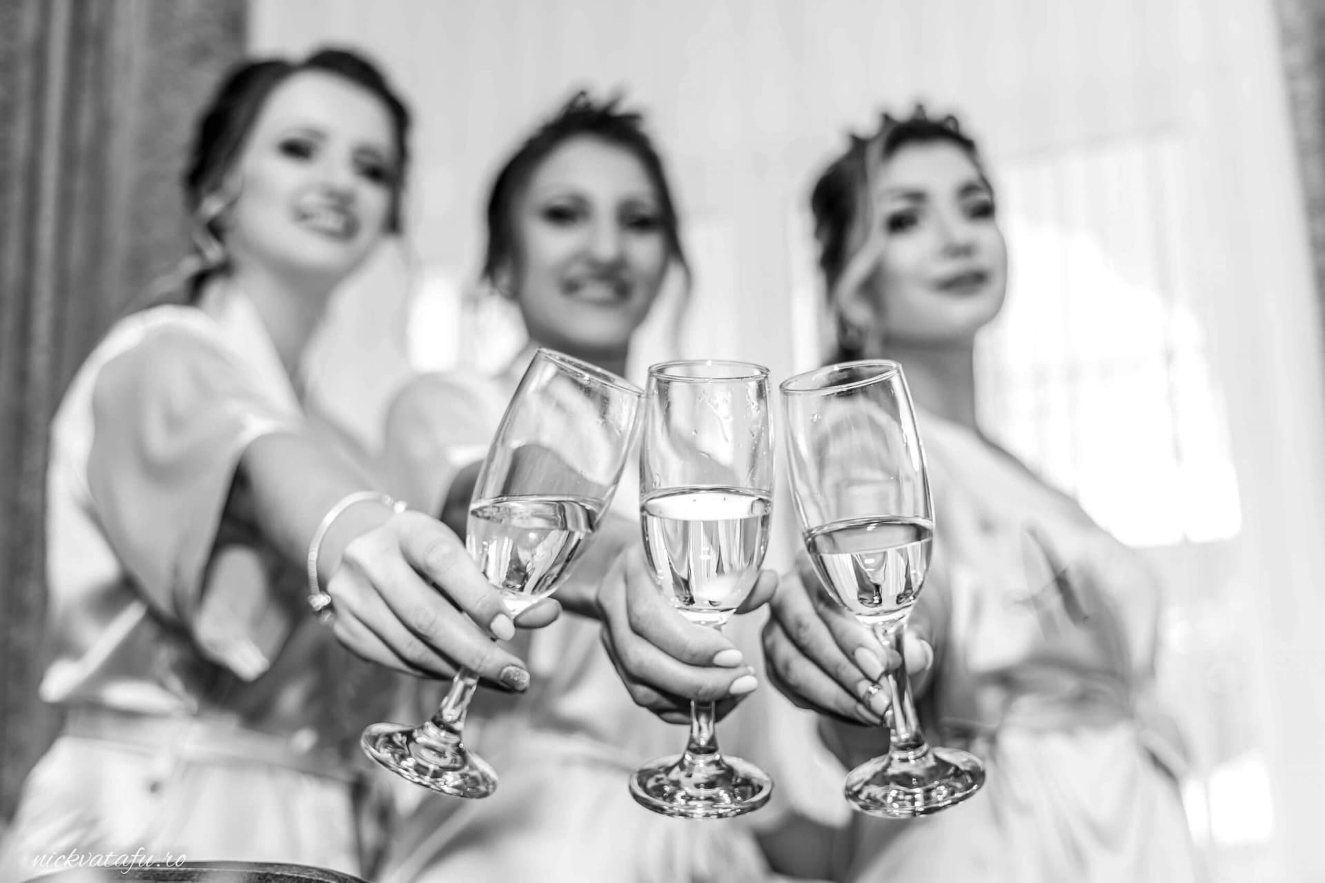 Trei domnișoare de onoare ridică cu bucurie paharele, pregătind atmosfera unei fotografii de nuntă pline de emoție.