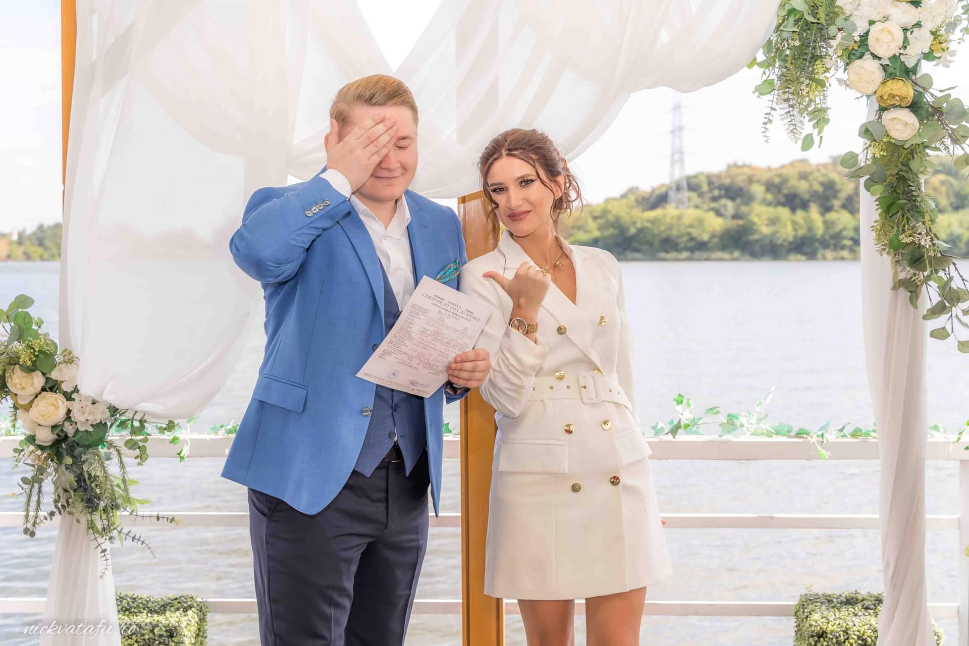 Mire și mireasă zâmbind emoționați în fața invitaților, la ceremonia de nuntă pe malul apei.