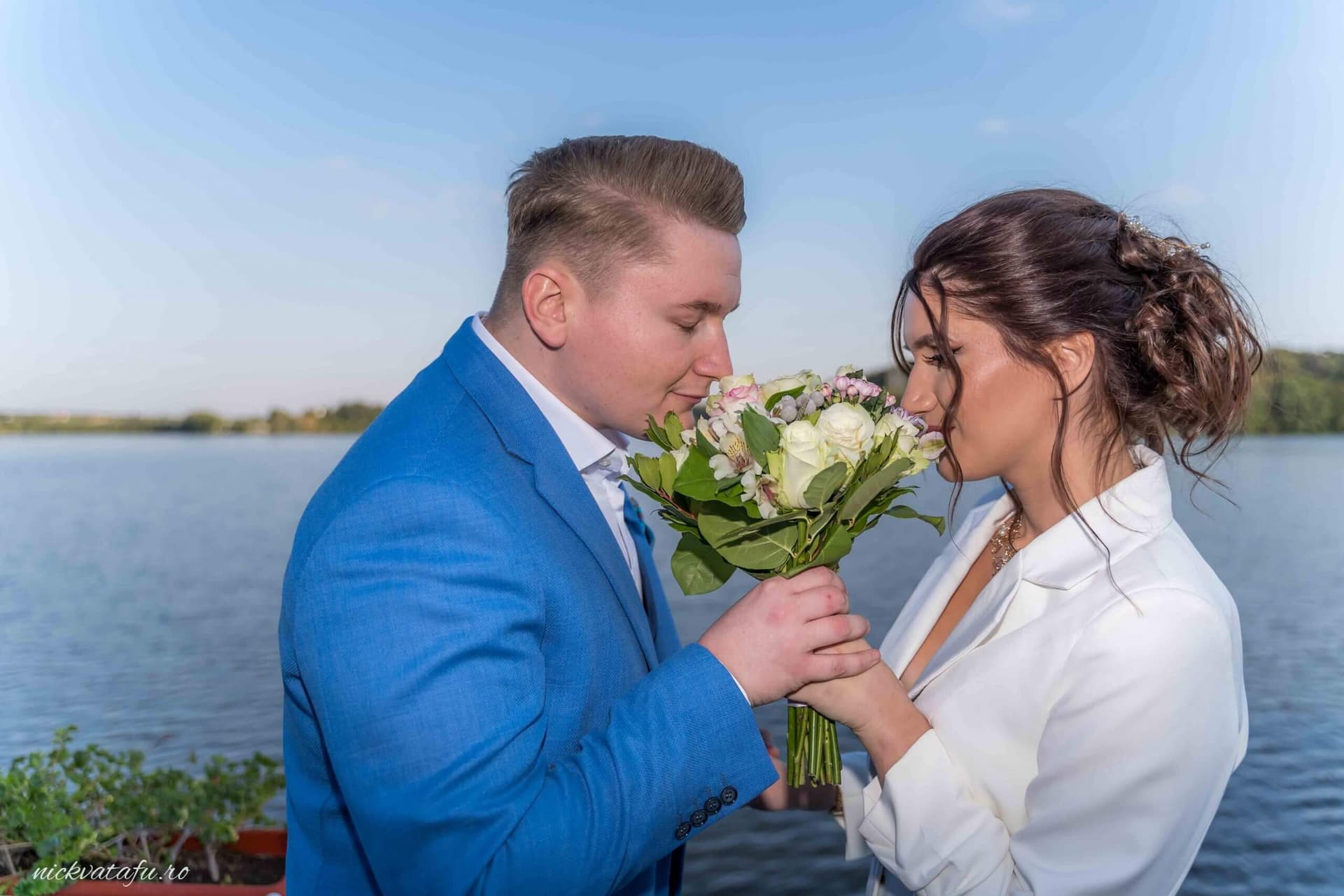 Mire și mireasă la malul apei, admirând buchetul de flori, fotografie de nuntă plină de emoție și romantism.