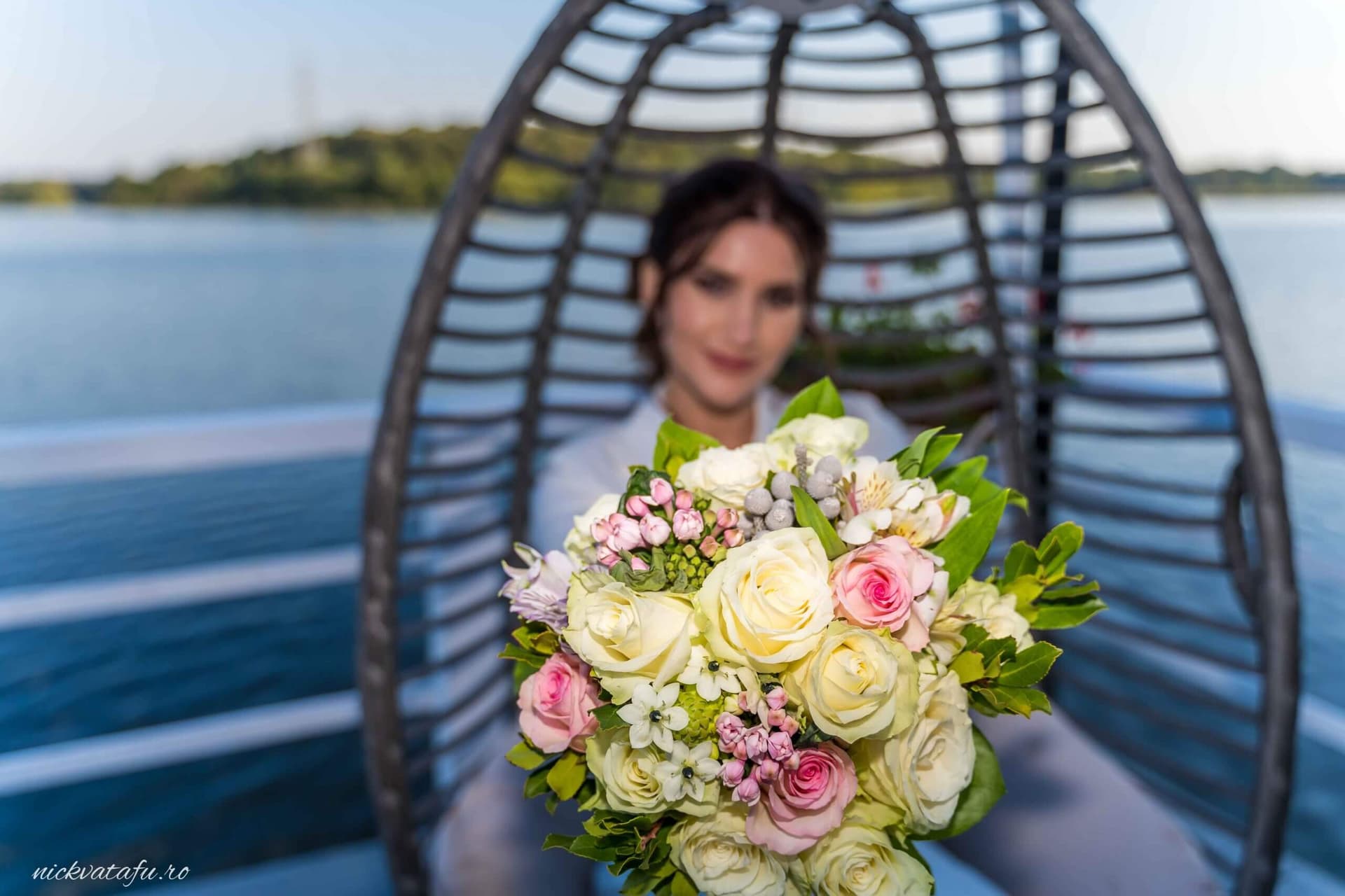 Mireasă zâmbitoare cu buchet de flori, în fundal un peisaj lacustru, fotografie elegantă de nuntă.