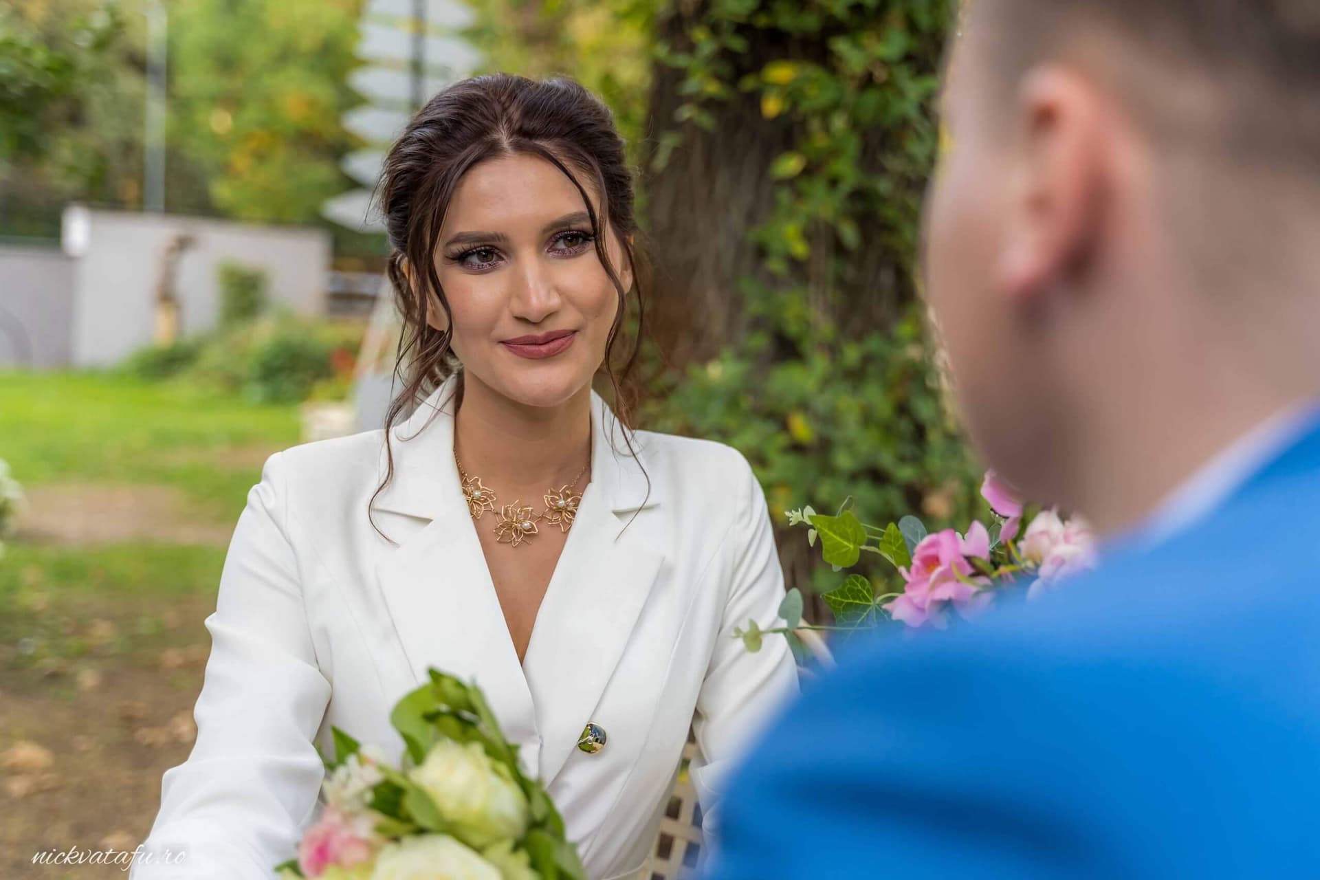 Mireasă zâmbitoare, cu buchet în mână, privind emoționată spre mire într-o grădină luxuriantă, fotografie nuntă.