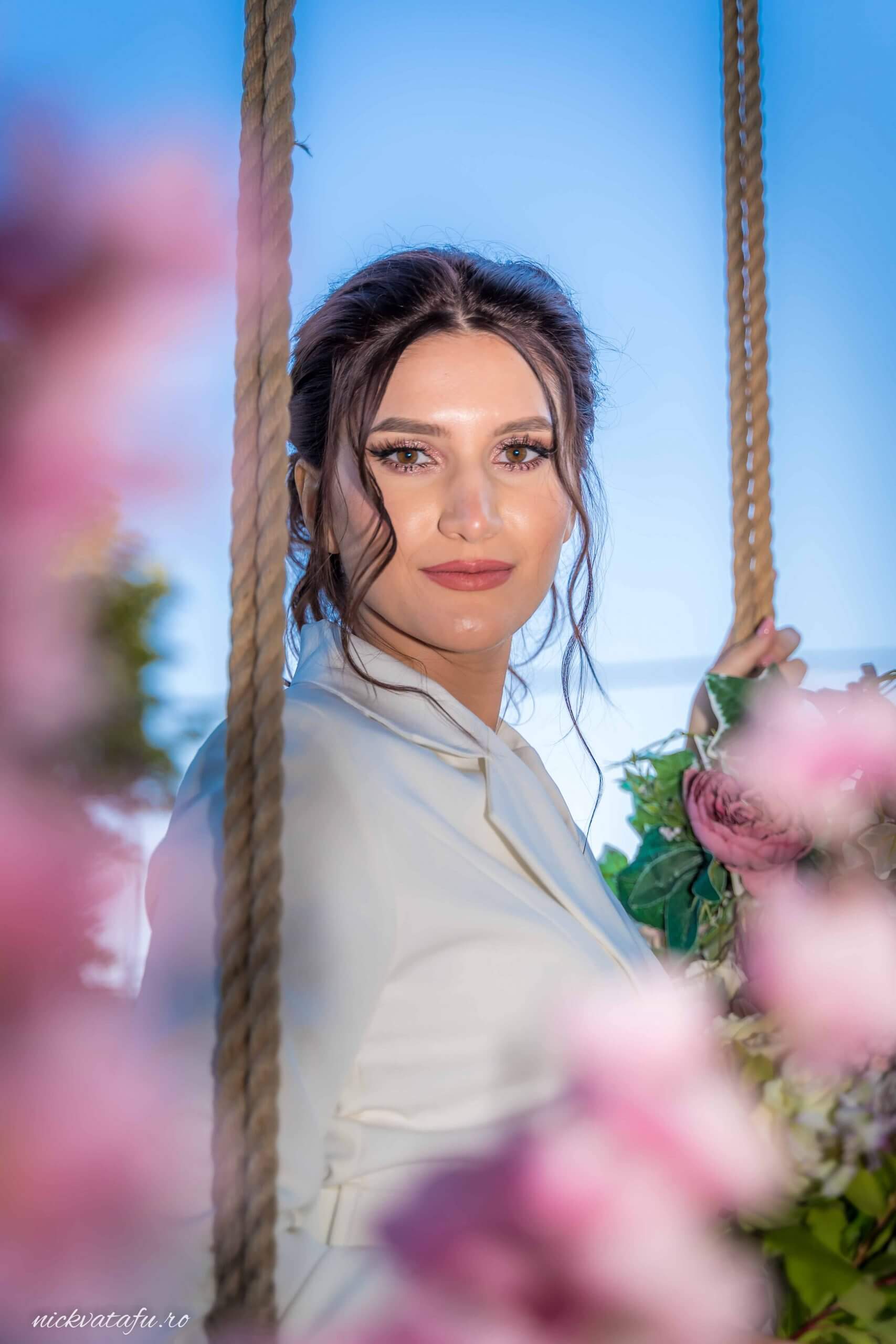 Mireasă zâmbitoare pe o leagăn floral, emanând emoție și eleganță, fotografie de nuntă în natură.