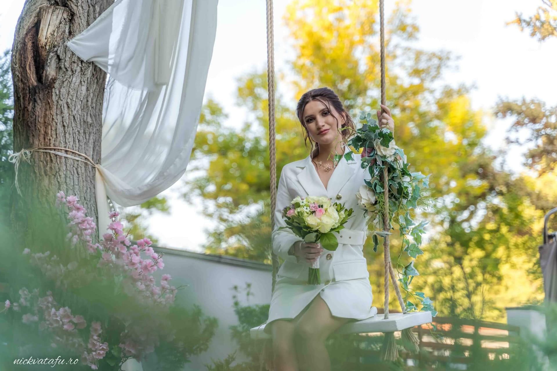 Mireasă zâmbitoare pe leagăn, cu buchet de flori, în mijlocul unui decor natural vibrant, fotografie de nuntă elegantă.