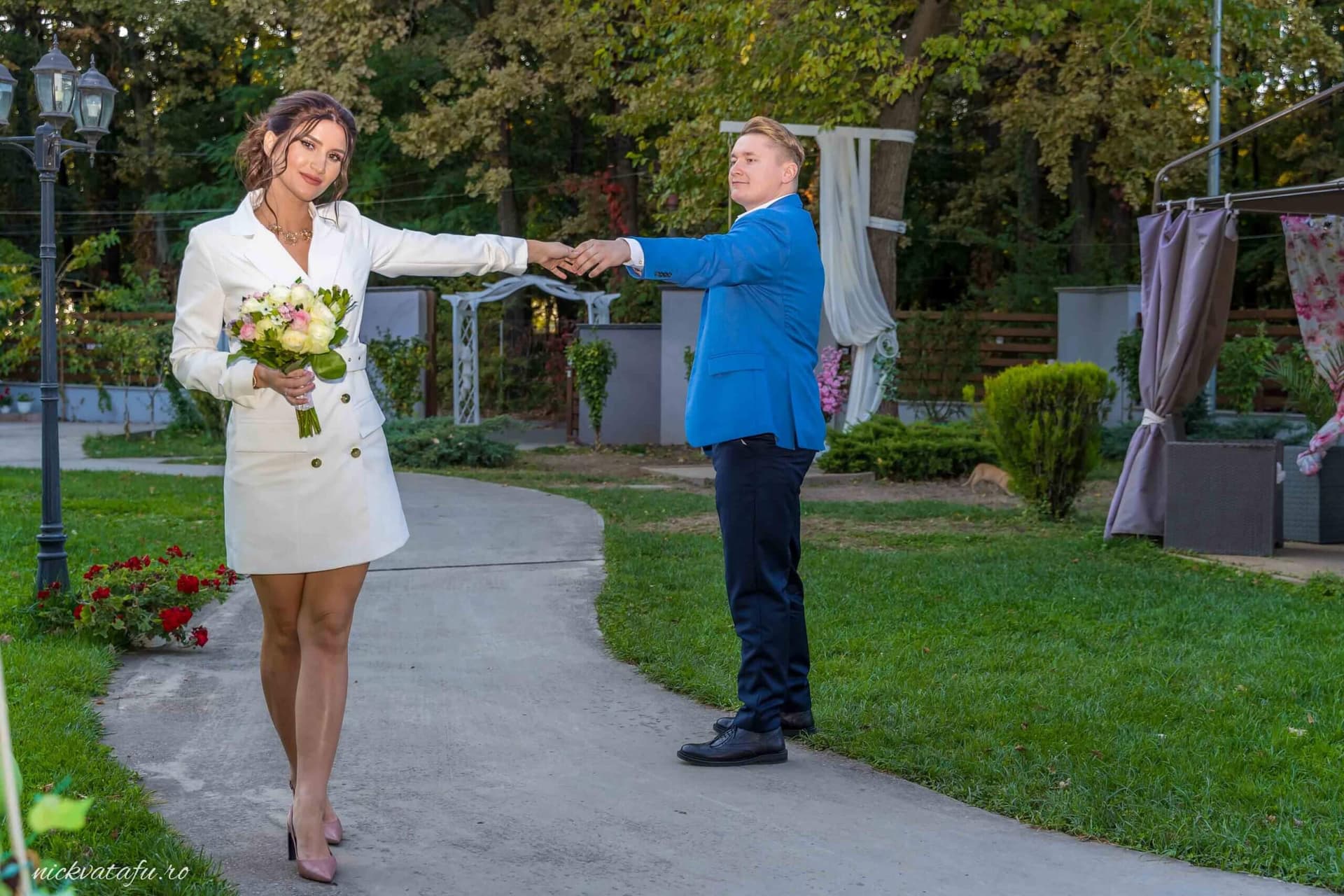 Mireasă zâmbitoare și mire îmbrăcat elegant, plimbându-se pe aleea unei grădini, fotografie nuntă plină de emoție.