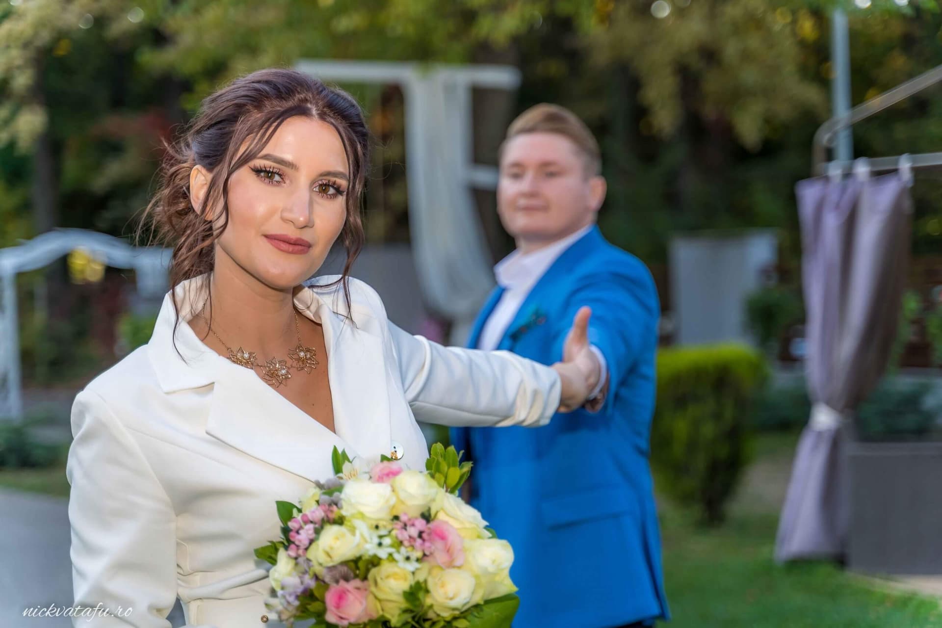 Mireasă zâmbitoare cu buchet de flori, iar mirele o urmărește cu admirație, fotografie de nuntă plină de emoție.