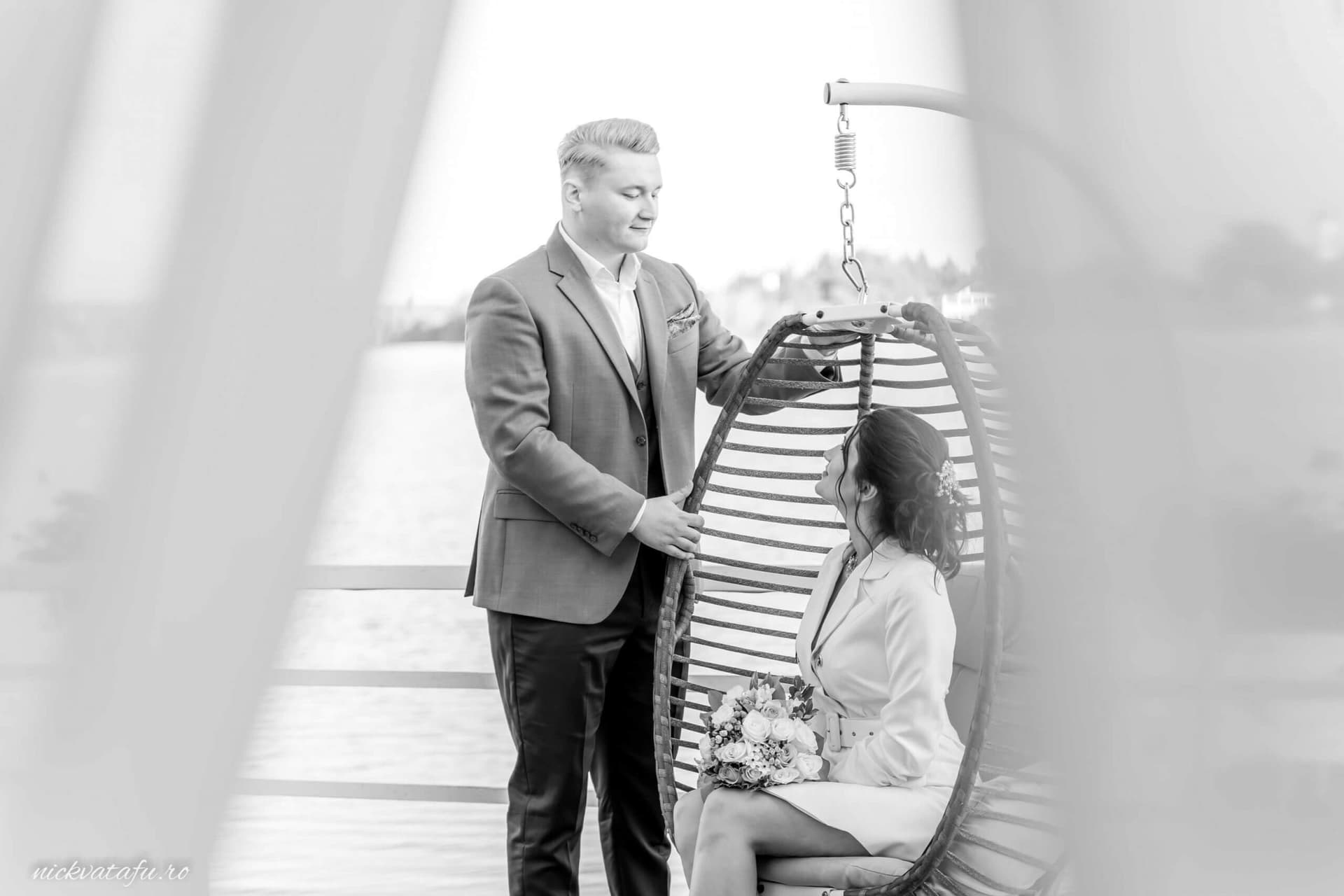 Mire și mireasă zâmbind și interacționând pe un leagăn, fotografie elegantă de nuntă la malul apei.