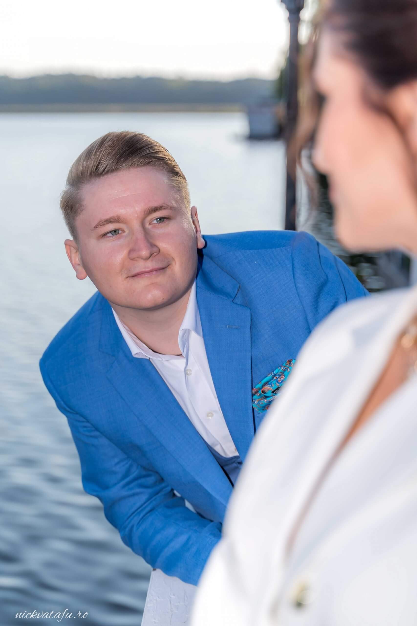 Bărbat îmbrăcat elegant, zâmbind către o persoană, pe fundalul apei, fotografie nuntă plină de emoție.