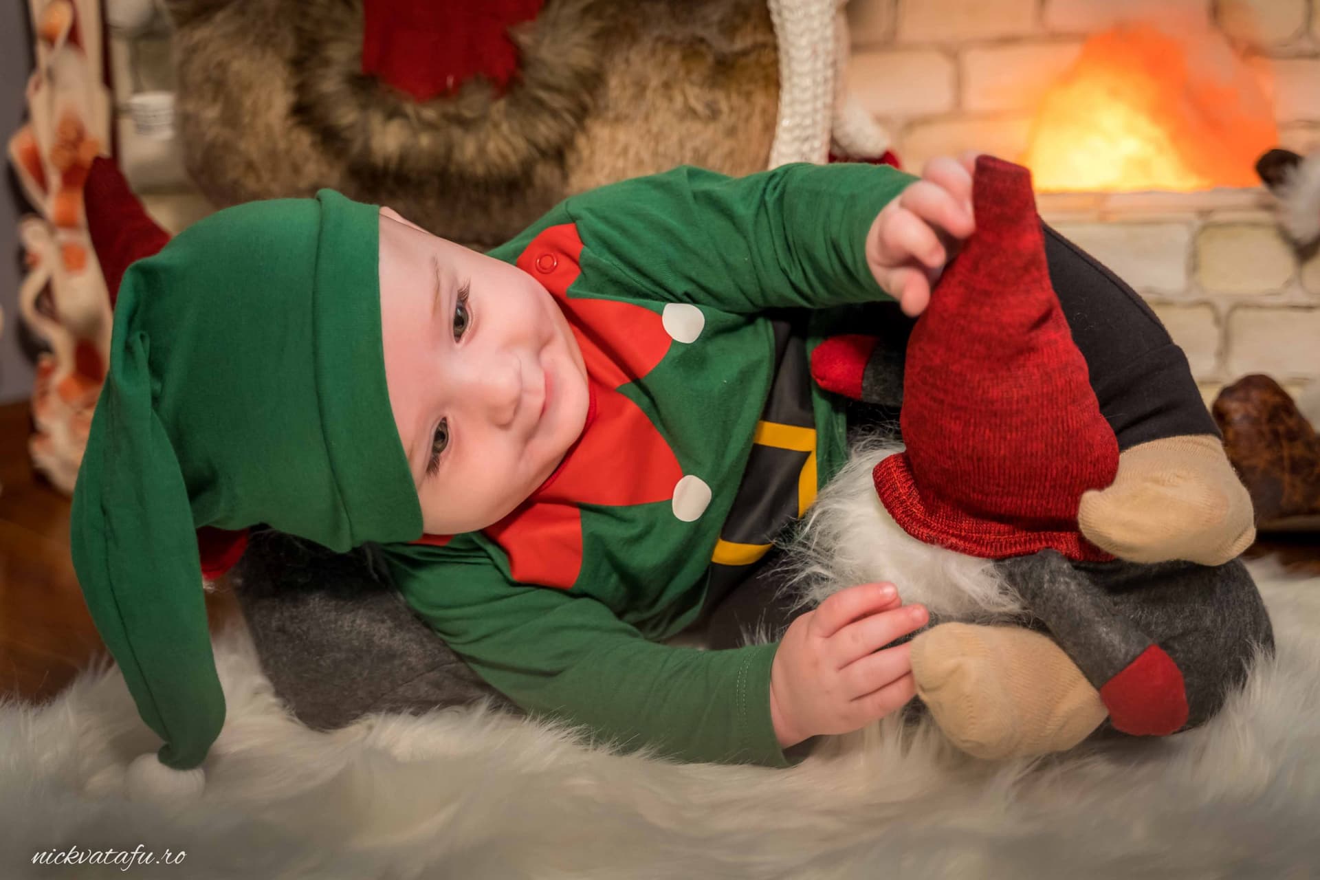 Băiețel îmbrăcat în costum de elf, zâmbind și jucându-se cu jucării, atmosferă festivă de Crăciun.
