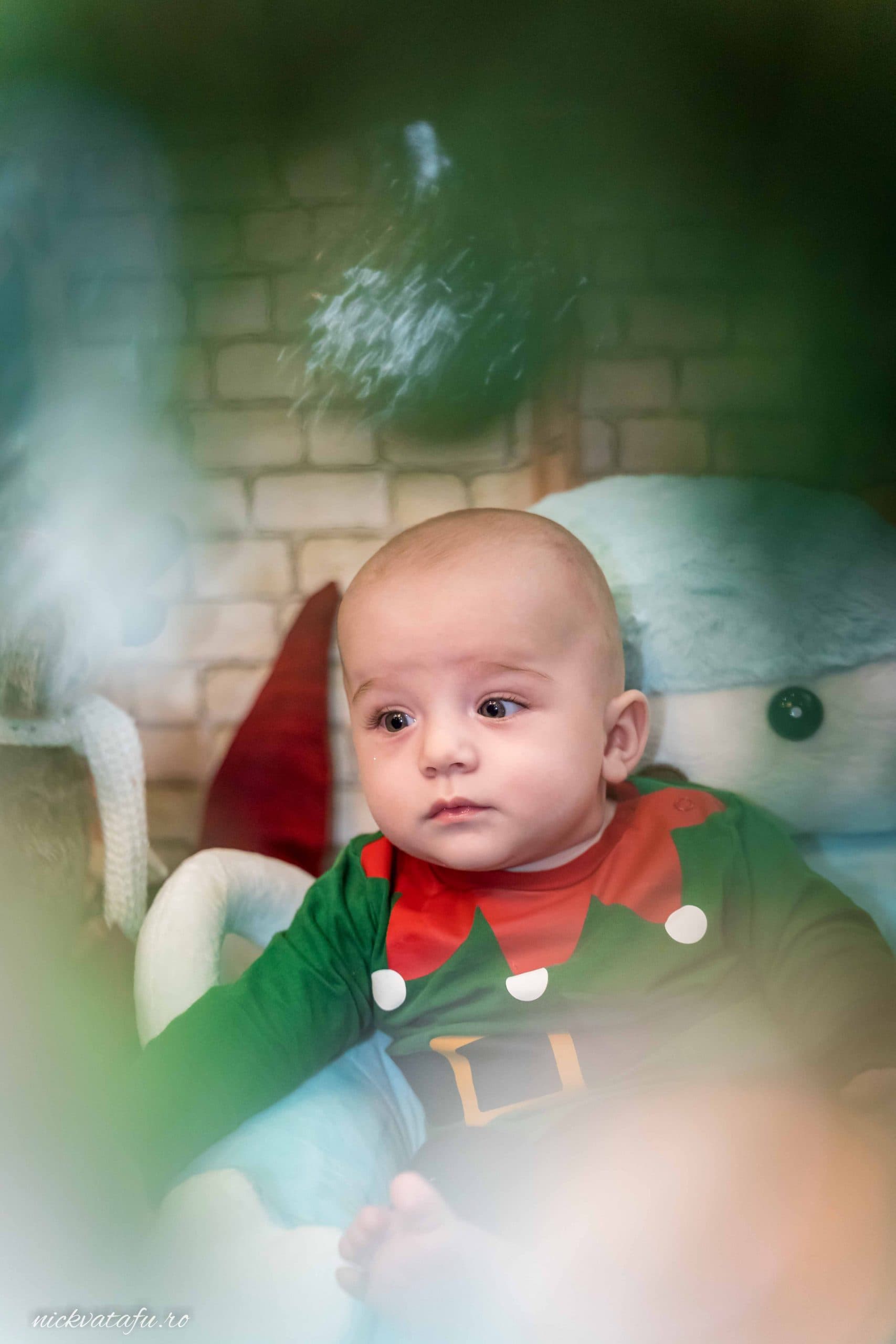 Bebeluș îmbrăcat în costum de elf, privind cu curiozitate, într-un decor festiv de sărbătoare.