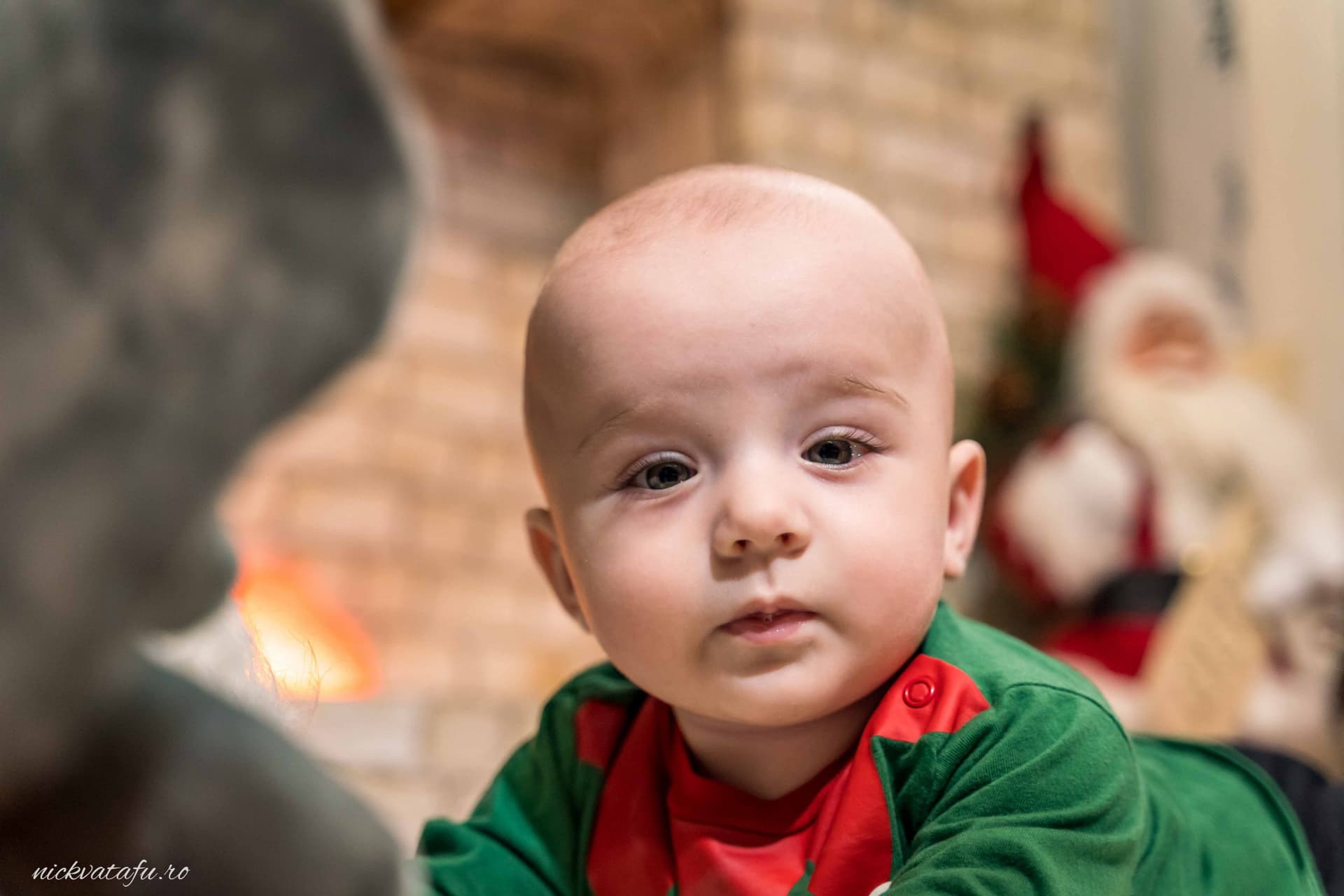 Bebeluș cu privire curioasă, îmbrăcat în haine festive, într-un decor de Crăciun plin de emoție și căldură.