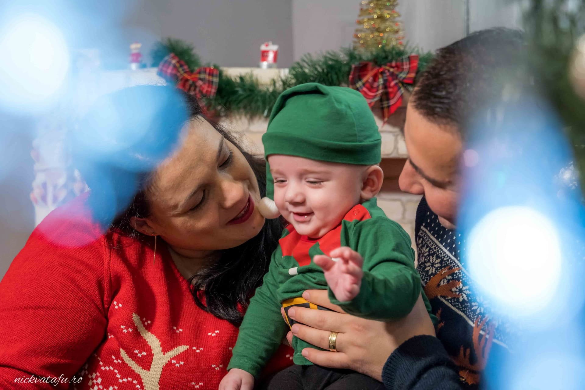 Familie fericită sărbătorind Crăciunul, cu un bebeluș zâmbitor îmbrăcat în verde, fotografiat într-un decor festiv.