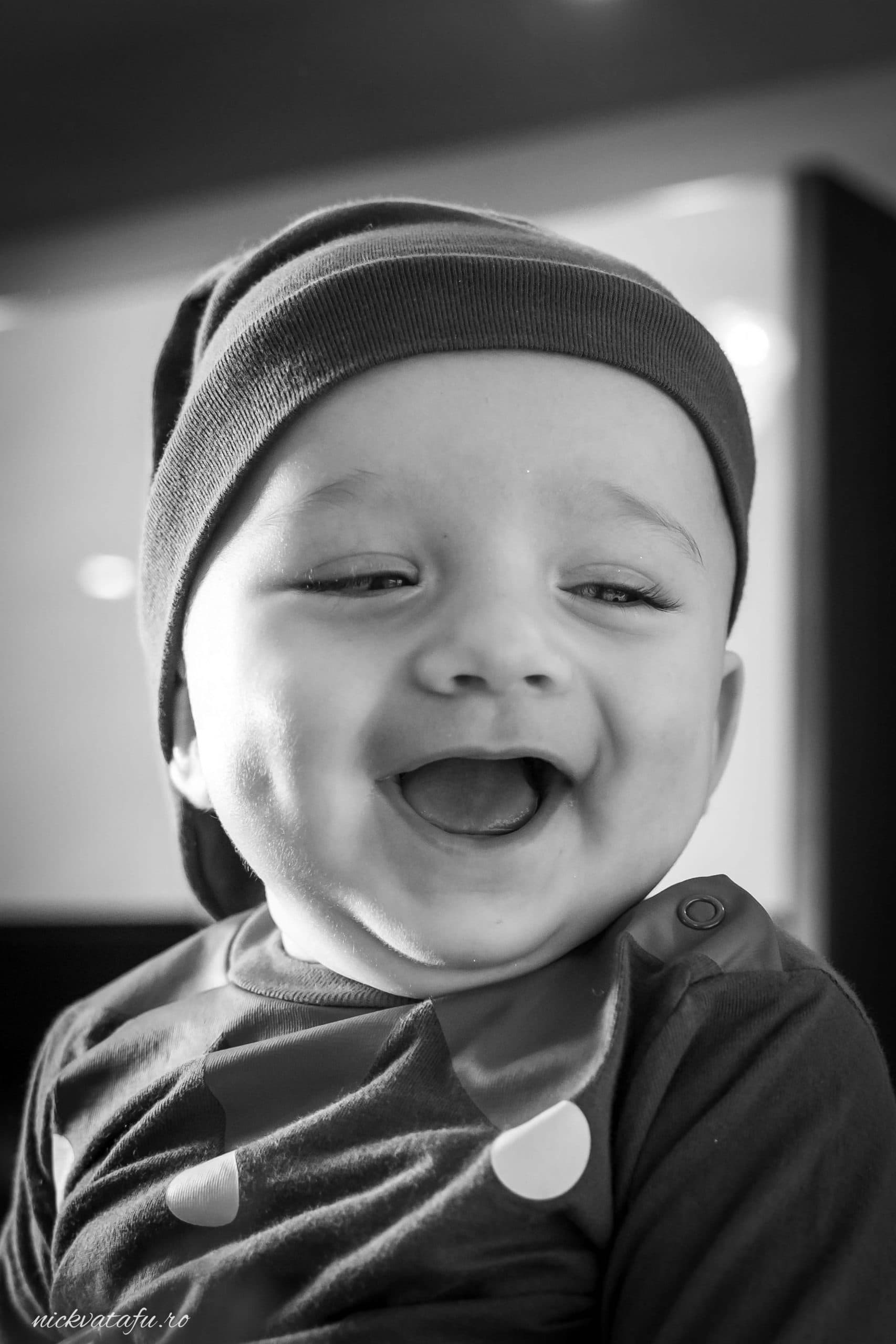 Bebeluș zâmbitor cu obraje rumeni, purtând o căciulă, emoție pură într-o fotografie de botez.