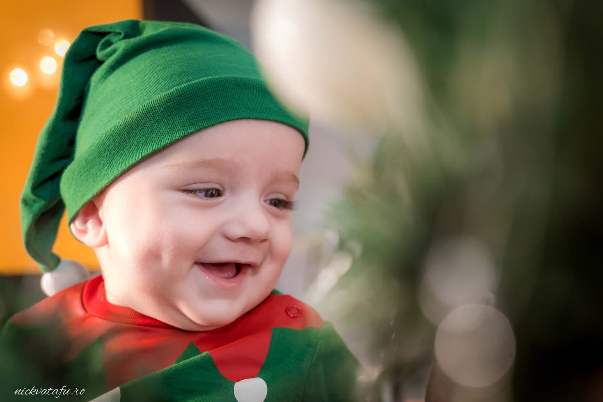 Băiețel vesel îmbrăcat în costum de elf, zâmbind lângă un brad de Crăciun, fotografiat într-un moment plin de emoție.